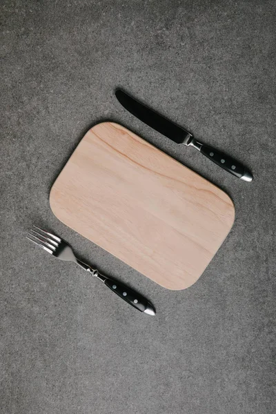 Top view of wooden board with fork and knife on grey — Stock Photo
