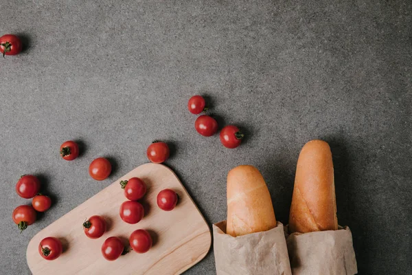 Top view of baguettes in paper bags, fresh tomatoes and wooden board on grey — Stock Photo