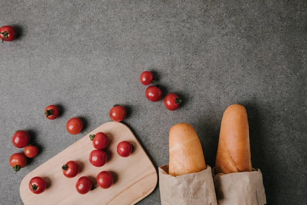 Vista superior de baguettes en bolsas de papel, tomates frescos y tablero de madera en gris - foto de stock