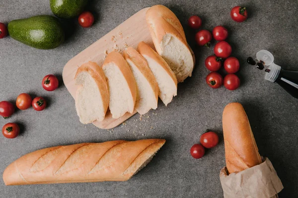 Top view of sliced baguette on wooden board, fresh tomatoes and avocados on grey — Stock Photo