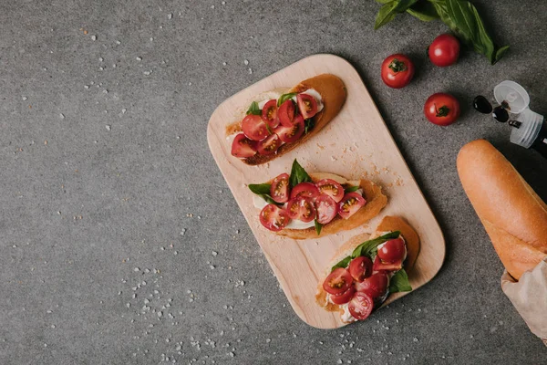 Blick von oben auf leckere Tomaten-Bruschetta auf Holzbrett und frische Zutaten auf Grau — Stockfoto