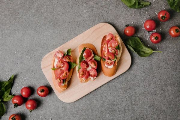 Vista superior de deliciosa bruschetta em tábua de madeira e ingredientes frescos em cinza — Fotografia de Stock