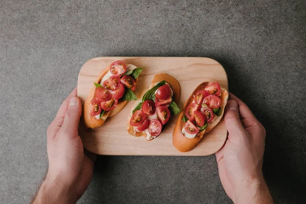 Cropped shot of person holding wooden board with delicious tomato bruschetta — Stock Photo
