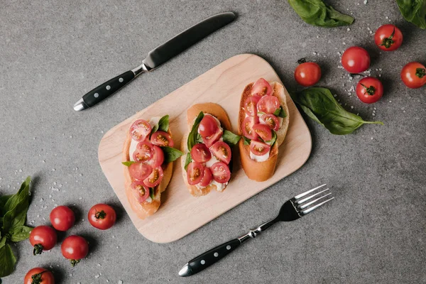 Vue de dessus de savoureuse bruschetta italienne sur planche de bois, ingrédients et couverts sur gris — Photo de stock