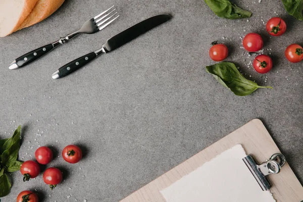 Vista superior de tenedor y cuchillo, portapapeles en blanco, baguette, albahaca y tomates frescos en gris - foto de stock