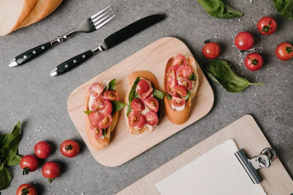 Draufsicht auf traditionelle italienische Bruschetta auf Holzbrett, Zutaten, Besteck und Klemmbrett auf grau — Stockfoto