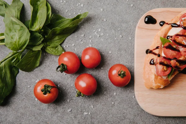 Blick von oben auf köstliche Bruschetta auf Holzbrett, Basilikum und frische Tomaten auf Grau — Stockfoto