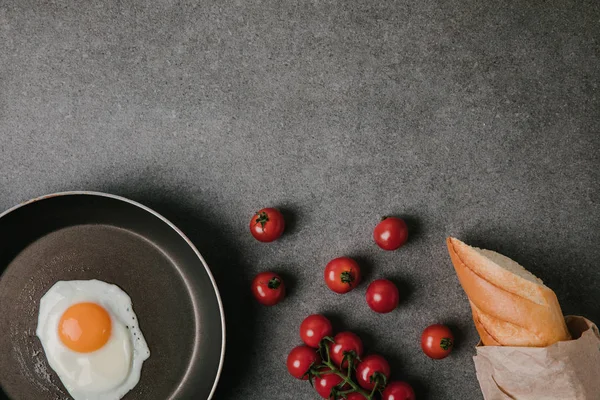 Верхний вид жареного яйца на сковороде, свежие помидоры и багет в бумажном пакете на сером — стоковое фото
