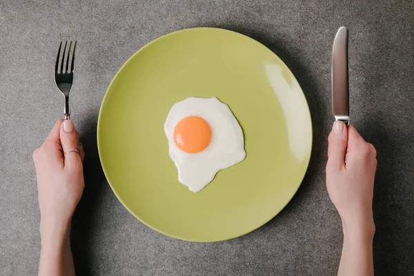 Tiro recortado de la persona sosteniendo tenedor y cuchillo listo para comer huevo frito sabroso en el plato - foto de stock