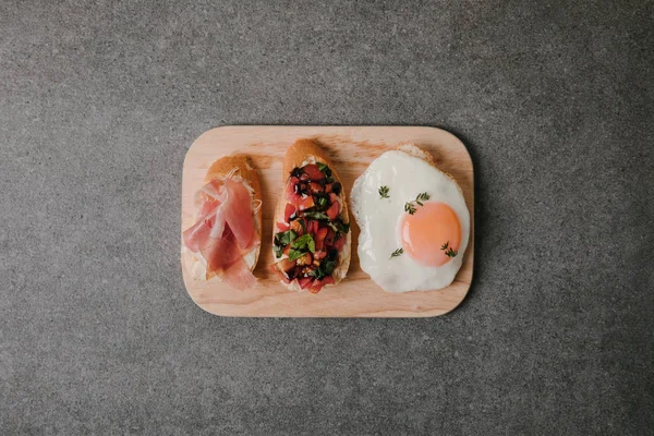 Draufsicht auf köstliche Antipasto Bruschetta mit Spiegelei auf Holzbrett auf grau — Stockfoto