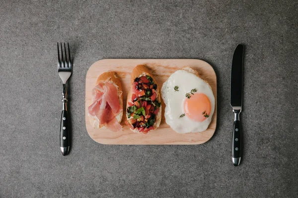 Vue de dessus de bruschetta antipasto gastronomique avec oeuf frit et fourchette avec couteau sur gris — Photo de stock