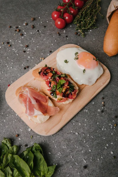 Delicious antipasto bruschetta and fried egg on wooden cutting board and ingredients on grey — Stock Photo