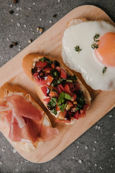 Delicious antipasto bruschetta and fried egg on wooden cutting board and spices on grey — Stock Photo