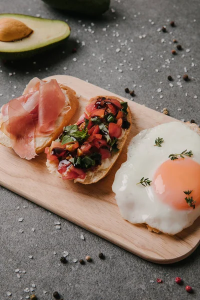 Close-up view of delicious antipasto bruschetta and fried egg on wooden cutting board — Stock Photo