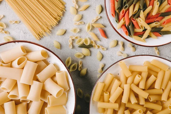 Draufsicht auf verschiedene rohe Pasta in Schüsseln auf chaotischer Betonoberfläche — Stockfoto