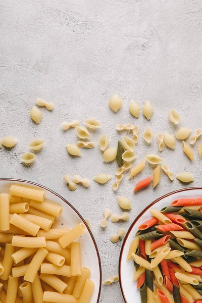 Draufsicht auf verschiedene rohe Pasta in Schüsseln auf chaotischer Betontischplatte — Stockfoto