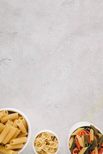 Draufsicht auf verschiedene Arten roher Pasta in Schüsseln auf Betontischplatte — Stockfoto