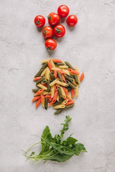 Top view of raw colorful pasta with tomatoes and arugula on concrete surface — Stock Photo
