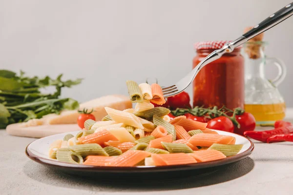 Garfo com macarrão colorido perfurado acima da placa em mesa de concreto com vários alimentos no fundo — Fotografia de Stock