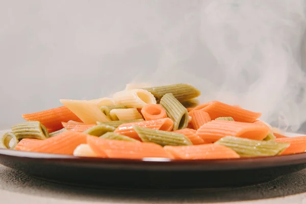 Nahaufnahme bunter Penne-Pasta, die auf einem Teller auf einer Betonplatte dampft — Stockfoto