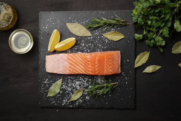 Piece of salmon with salt on black table with lemon and herbs — Stock Photo