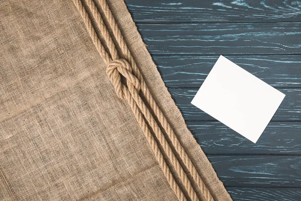 Top view of knotted brown nautical rope on sackcloth and empty paper on wooden surface — Stock Photo