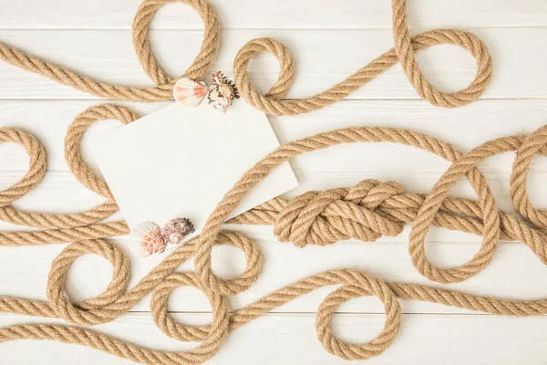 Draufsicht auf leeres Papier mit Muscheln auf braunen nautisch geknüpften Seilen auf weißer Holzoberfläche — Stockfoto