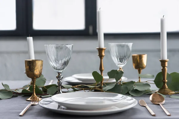 Vista de cerca de la mesa rústica con copas de vino, eucalipto, cubiertos vintage, velas en candelabros y platos vacíos - foto de stock