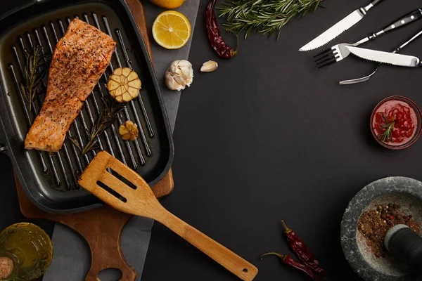 Top view of arranged grilled salmon steak on wooden cutting board, ingredients, sauce and cutlery on black tabletop — Stock Photo