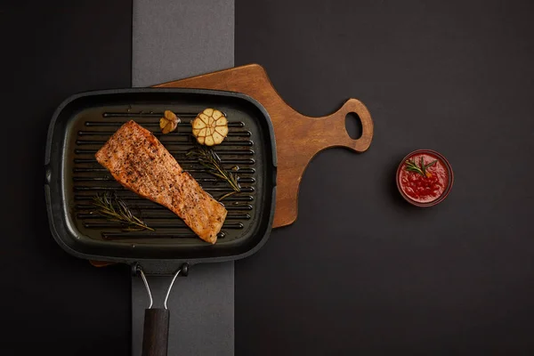 Top view of arranged grilled salmon steak on wooden cutting board and sauce on black tabletop — Stock Photo