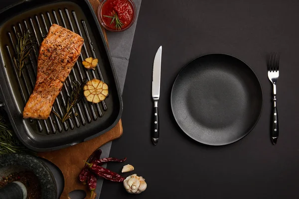 Top view of arranged grilled salmon steak on wooden cutting board, sauce and cutlery on black tabletop — Stock Photo