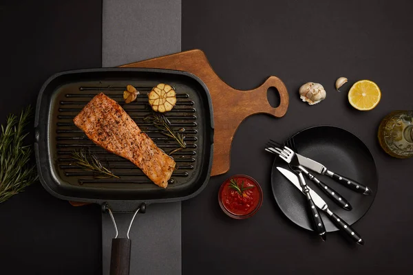 Vue de dessus du steak de saumon grillé arrangé sur planche à découper en bois, sauce et couverts sur plateau noir — Photo de stock
