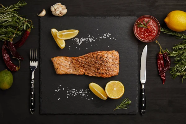Top view of grilled salmon steak with pieces of lemon, arranged ingredients around and cutlery on black surface — Stock Photo