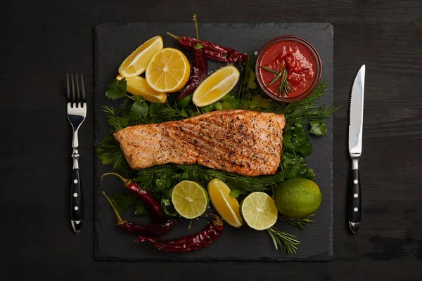 Top view of grilled salmon steak, pieces of lime and lemon, chili peppers, sauce and cutlery on black surface — Stock Photo