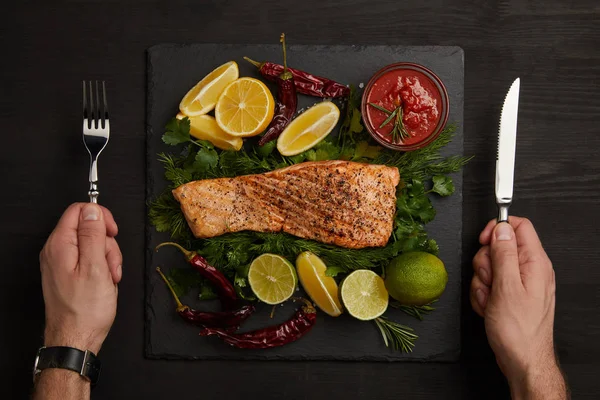 Partial view of male hands with cutlery, grilled salmon steak, sauce and arranged citrus fruits on black surface — Stock Photo