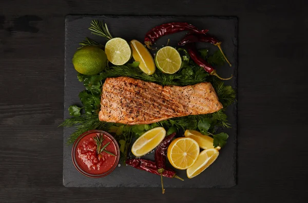 Top view of grilled salmon steak, pieces of lime and lemon, chili peppers and sauce on black surface — Stock Photo