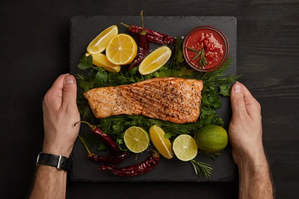 Partial view of male hands, grilled salmon steak, sauce and arranged citrus fruits on black surface — Stock Photo