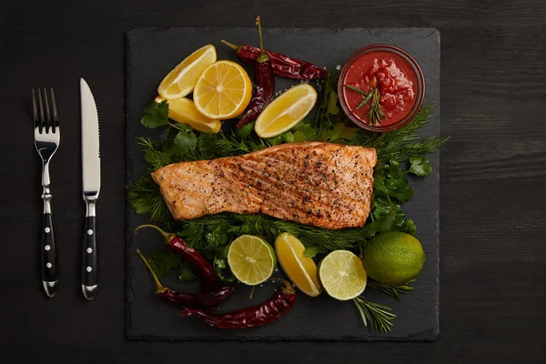 Vista dall'alto di bistecca di salmone alla griglia, pezzi di lime e limone, salsa e posate sulla superficie nera — Foto stock