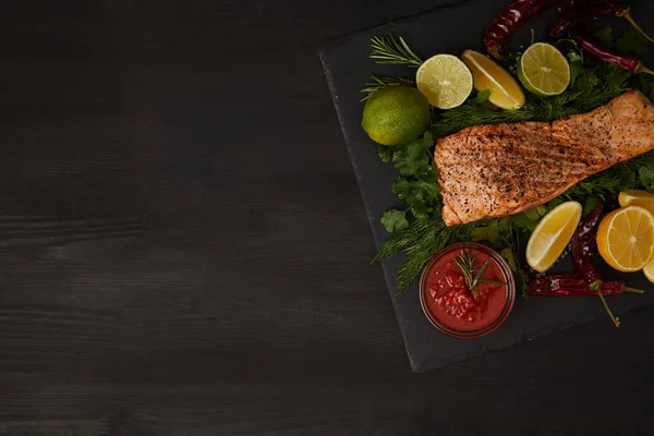 Vue du dessus du steak de saumon grillé, morceaux de citron vert et de sauce à la surface noire — Photo de stock