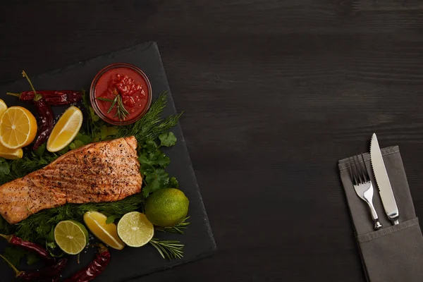 Top view of grilled salmon steak, pieces of lime and lemon, sauce and cutlery on black surface — Stock Photo