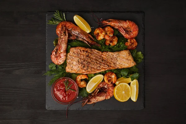 Vue du dessus du steak de saumon grillé, crevettes, morceaux de citron et sauce sur la surface noire — Photo de stock