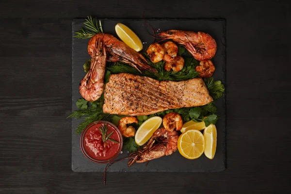 Vista superior del filete de salmón a la parrilla, camarones, trozos de limón y salsa en la superficie negra - foto de stock