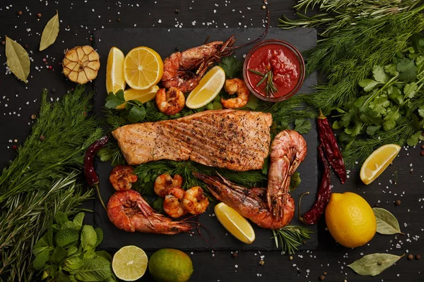 Vista dall'alto di bistecca di salmone alla griglia, gamberetti, pezzi di limone, salsa e spezie sulla superficie nera — Foto stock