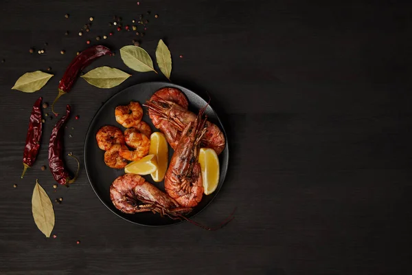 Top view of grilled shrimps and lemon pieces on plate with arranged spices around on black surface — Stock Photo