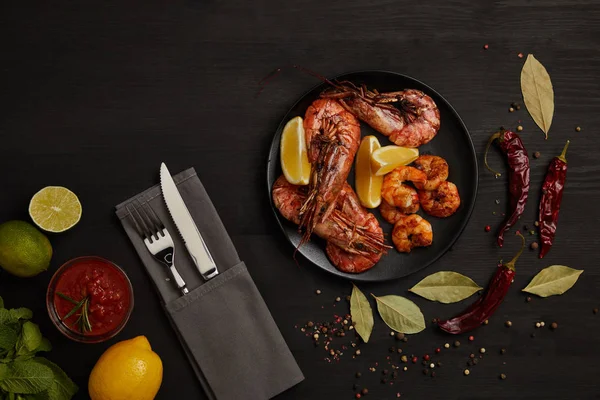 Flat lay with grilled shrimps with lemon pieces on plate and arranged sauce, spices, ingredients and cutlery around on black surface — Stock Photo