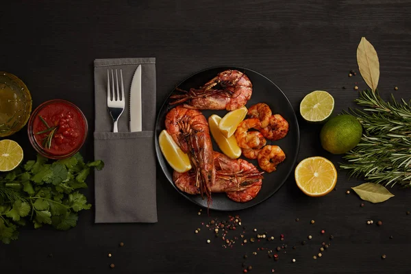 Flat lay com camarões grelhados com pedaços de limão na placa e molho arranjado, especiarias, ingredientes e talheres em torno de superfície preta — Fotografia de Stock