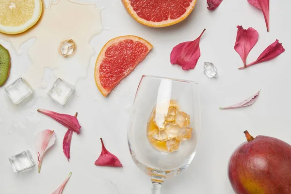 Ansicht der Anordnung von Weinglas, exotischen Früchten, Eiswürfeln und Blütenblättern auf weißer Oberfläche — Stockfoto