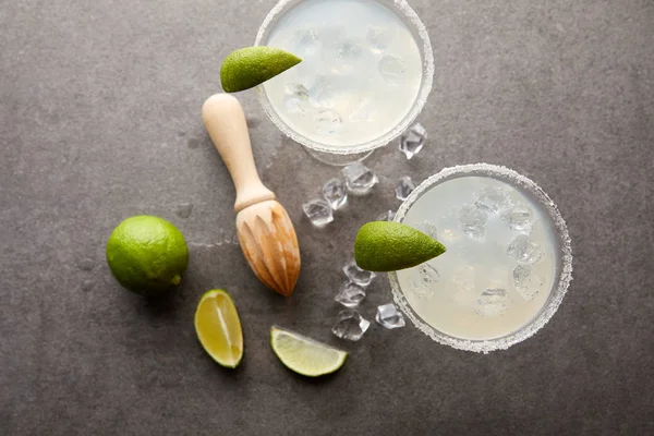 Posa piatta con cocktail margarita con pezzi di lime, cubetti di ghiaccio e spremiagrumi in legno su tavolo grigio — Foto stock