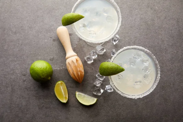 Posición plana con cócteles de margarita con trozos de lima, cubitos de hielo y exprimidor de madera en la mesa gris - foto de stock