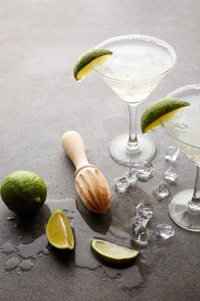 Close up view of alcohol cocktails with pieces of lime, ice cubes and wooden squeezer on grey tabletop — Stock Photo